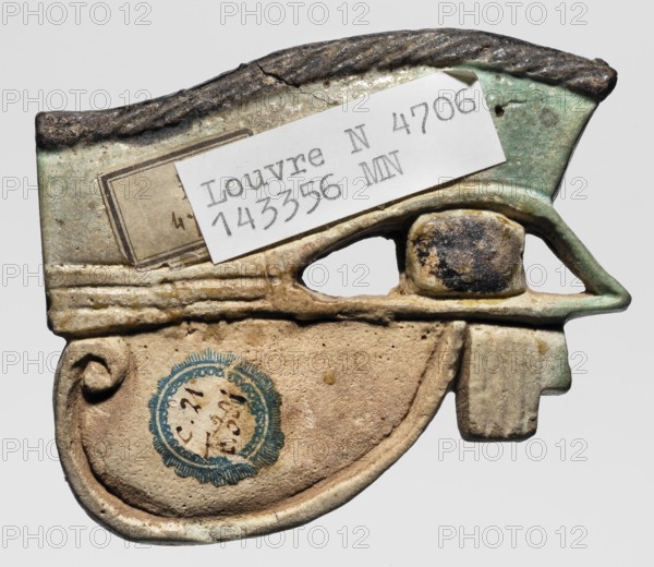 Amulet - Udjat Eye, Udjat eye designed as a flat plaque with details of the eye marked with a fairly wide engraved line. Eyebrow and pupil painted black. A pin placed above the eyebrow. ca. 747–656 BC; 25th Dynasty, Egypt, grave goods, amulet body part, height 5.5 cm, width 7 cm, depth 1 cm, The image shows a worn, irregularly shaped object with a combination of smooth and rough textures, primarily in shades of beige, tan, pale green, and dark brown, with areas of blackened discoloration; a group of small paper labels rest on the upper surface, and there is a round blue circular stamp-like mark on the lower beige area; the object has a curved lower portion, a hollowed central opening, and a protruding rectangular element on the right side.