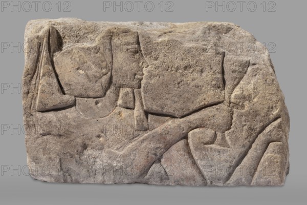 Relief depicting Ramesses II with a ritual vessel, uraeus iconographic, ibes wig iconographic, ritual vessels iconographic, king iconographic. The king is depicted in right profile. Preserved face and part of a wig with a hanging uraeus. Around the neck is a wide, smooth necklace. In the hands are a hes vase and a possible incense burner not preserved. Inscription: Ramesses II ruler of Egypt; ca. 1279–1213 BC. Polish excavations in Faras Sudan provenance. Scen. Gal. Eg., religious scenes, sacred architecture, Egyptian architecture. c. 1279–1213 BC; New Kingdom, 19th Dynasty reign of Ramesses II. Found at Faras; Aksha site. Architectural element, bas-relief. Height 33.5 cm, width 54 cm, depth 23 cm, weight 49.5 kg., A weathered stone block with carved relief forms a rectangular shape against a plain gray background, showing a profile of a human face and portions of a body in shallow bas-relief. The stone surface is textured and rough with worn edges and shallow grooves; the carving outl...