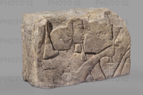 Relief depicting Ramesses II with a ritual vessel; uraeus on wig; hes vase and possible incense burner not preserved; king shown in right profile with preserved face and part of wig; wide smooth necklace around the neck; ibes wig iconographic; ritual vessels iconographic; king iconographic; religious scenes; sacred and Egyptian architecture; architectural element, bas-relief; maker, Ramesses II ruler of Egypt; ca. 1279–1213 BC, New Kingdom, 19th Dynasty reign of Ramesses II; provenance: Faras; findspot: Aksha; c. 1279–1213 BC; height 33.5 cm, width 54 cm, depth 23 cm, weight 49.5 kg., A roughly rectangular carved stone block with a weathered surface showing a carved profile of a human figure and surrounding abstract shapes; the stone appears in shades of light brown, beige, tan, gray, and hints of muted reddish tones, with textured rough and smoother worn areas, visible carved lines and shallow relief, and a neutral gray background.