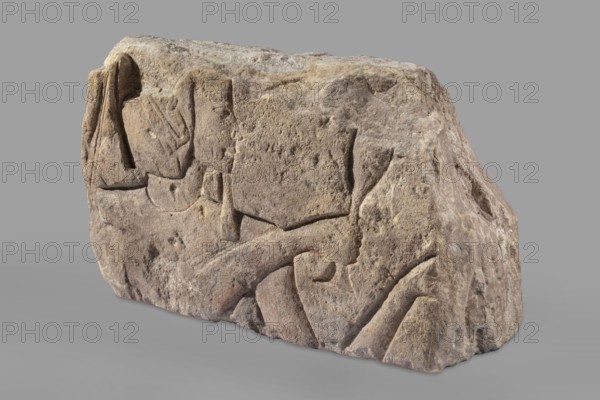 Relief depicting Ramesses II with a ritual vessel, uraeus iconographic, ibes wig iconographic, ritual vessels iconographic, king iconographic; The king is depicted in right profile. Preserved face and part of a wig with a hanging uraeus. Around the neck is a wide, smooth necklace. In the hands are a hes vase and a possible incense burner not preserved. Ramesses II ruler of Egypt; ca. 1279–1213 BC, c. 1279–1213 BC; New Kingdom, 19th Dynasty reign of Ramesses II. Polish excavations in Faras Sudan, Faras; Aksha. Architectural element, bas-relief. Height 33.5 cm, width 54 cm, depth 23 cm, weight 49.5 kg., A roughly rectangular carved stone block with an uneven, weathered surface sits against a plain grey background; the stone shows a textured, pitted surface with carved recesses and raised areas forming abstract, angular and curved shapes. The block's colors include various shades of grey, beige, tan, and hints of brown and muted yellow, with slightly darker shadowed crevices and some subt...