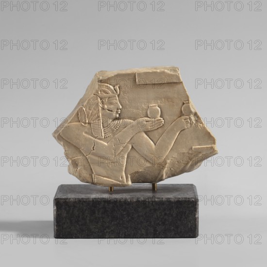 Relief depicting King Amasis - copy of artifact no. 141276, contemporary product, copy, height 7.1 cm, width 9.2 cm, A carved pale beige stone fragment mounted on a dark gray rectangular base shows a raised relief of a human profile wearing a headdress and a detailed collar, with an arm extended holding a rounded object and another arm raised holding a similar rounded object; the stone has visible cracks and chipped edges and the scene is set against a smooth neutral gray background with soft shadows.