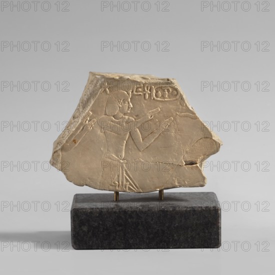 Relief depicting King Amasis — copy of artifact no. 141276, creator unknown, contemporary product, place of origin unknown, copy, height 7.1 cm, width 9.2 cm, A pale beige stone fragment with carved shallow relief depicting a profile of a person facing right, mounted on a dark gray speckled rectangular base with two small brass supports; the carved figure shows a torso, arms raised with hands extended, a stylized short hairstyle, and a pleated skirt detail, with additional carved lines and shapes around the figure and along the fragment edges, all set against a neutral light gray background.