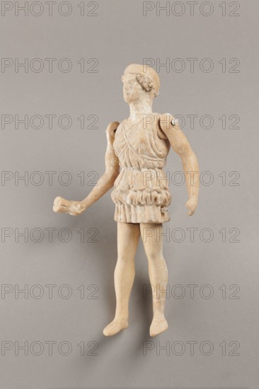 Doll with movable limbs, terracotta, made of terracotta. The doll depicts a woman in a short, pleated chiton and a bonnet; in her right hand she holds an instrument resembling castanets. The figure's oval head, directed slightly upwards, is set on a long, rather thick neck compared to the rest of the body. Lush curls protrude from under the headdress. The figurine's movable limbs are long compared to the rest of the body. 4th century BC, Attica object reported to come from Italy. Figurine, toy. Height 18.9 cm, width 8 cm, depth 3.5 cm, weight 0.13 kg., A small weathered figurine of a standing human figure with articulated shoulders and separate arms, shown against a plain neutral background. The figure wears a textured short tunic with layered folds and a belt-like section at the waist, and has short hair styled close to the head. The left arm is extended outward with a slightly bent elbow and an open hand, while the right arm hangs downward with a relaxed hand. The legs are straight w...