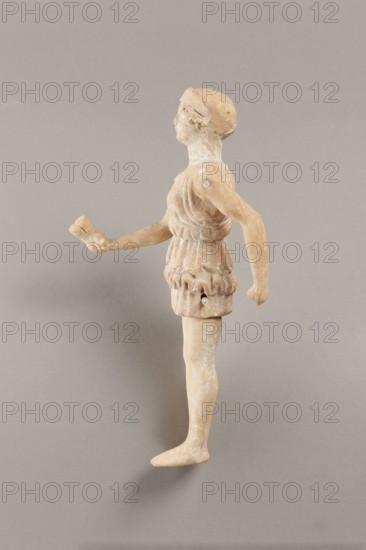 Doll with movable limbs, terracotta, dolls, Made of terracotta. The doll with movable limbs depicts a woman in a short, pleated chiton and a bonnet. In her right hand she holds an instrument resembling castanets. The figure's oval head, directed slightly upwards, is set on a long, rather thick neck compared to the rest of the body. Lush curls protrude from under the headdress. The figurine's movable limbs are long compared to the rest of the body. 4th century BC, Attica object possibly from Italy, figurine, toy, height 18.9 cm, width 8 cm, depth 3.5 cm, weight 0.13 kg., A small worn figurine of a person viewed in profile standing on a flat surface, made of a tan-beige material with patches of lighter whitish residue, wearing a short textured garment with folded and gathered details, one arm extended forward holding a small broken object and the other arm bent slightly back, a rounded head with simplified facial features and a cap-like headdress, visible joint connections at the shoulde...