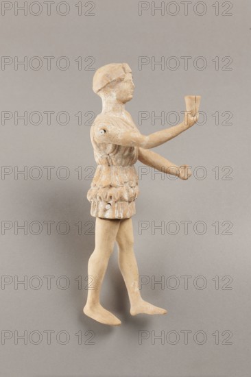 Doll with movable limbs, terracotta, dolls. Made of terracotta, the doll with movable limbs depicts a woman in a short, pleated chiton and a bonnet. In her right hand she holds an instrument resembling castanets. The figure's oval head, directed slightly upwards, is set on a long, rather thick neck compared to the rest of the body. Lush curls protrude from under the headdress. The figurine's movable limbs are long compared to the rest of the body. 4th century BC, Attica according to Weronika Krzemien the object comes from Italy. Figurine, toy. Height 18.9 cm, width 8 cm, depth 3.5 cm, weight 0.13 kg., A small standing figure made of a beige-tan material is shown against a neutral gray background, wearing a textured tunic with layered, ruffled details and a head covering; the figure has extended arms, one hand holding a small cup-shaped object, and a few small round attachments visible at shoulder and waist areas; the surface shows patches of lighter off-white traces and slight darker b...