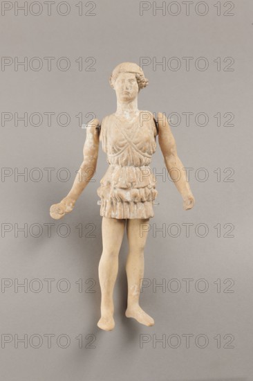 Doll with movable limbs, terracotta, dolls, Made of terracotta. The doll with movable limbs depicts a woman in a short, pleated chiton and a bonnet. In her right hand she holds an instrument resembling castanets. The figure's oval head, directed slightly upwards, is set on a long, rather thick neck compared to the rest of the body. Lush curls protrude from under the headdress. The figurine's movable limbs are long compared to the rest of the body. 4th century BC, Attica according to Weronika Krzemien the object comes from Italy. Figurine, toy. Height 18.9 cm, width 8 cm, depth 3.5 cm, weight 0.13 kg., A worn figurine of a human figure standing against a plain background, with a pale beige and light tan surface showing areas of lighter wear, wearing a short textured garment with layered ruffles, a fitted bodice with draped detailing, a rounded head with a simple hair-like shape, detached or partially detached arms held slightly away from the body and supported by small clear pegs, hands...