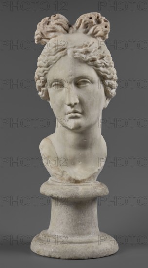 Head of the goddess Aphrodite, Roman workshop, early 1st century 1st century AD, sculpture; restitution Przeworsk; Aphrodite mythology; Roman sculpture; portraits; maker, A marble bust of a person displayed against a plain grey background, showing the head and upper neck on a rounded pedestal; the sculpture is off-white to light beige with subtle darker patches and weathered spots, the hair is carved in wavy locks gathered into a stylized arrangement at the top with textured, slightly darker areas, the facial features are smooth and slightly worn with closed or indistinct eyes, a straight nose, and a neutral mouth, the neck and shoulders show some surface pitting and small chips, and the pedestal base is cylindrical and matches the off-white and light grey tones of the bust.