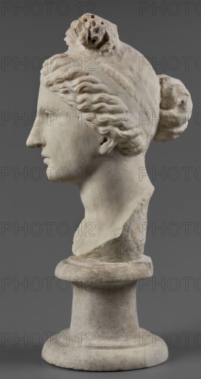 Head of the goddess Aphrodite, Roman workshop, restitution Przeworsk, Aphrodite mythology, Roman sculpture, portraits, early 1st century 1st century AD, sculpture, A carved stone bust of a woman shown in profile on a round pedestal, rendered in off-white and light beige tones with subtle darker beige and gray weathering; the woman has wavy hair pulled back into a bun and a textured hair ornament on top, with defined facial features including a straight nose, closed lips, and a visible ear; the surface shows small chips and rough patches, and the pedestal and background are shades of gray.