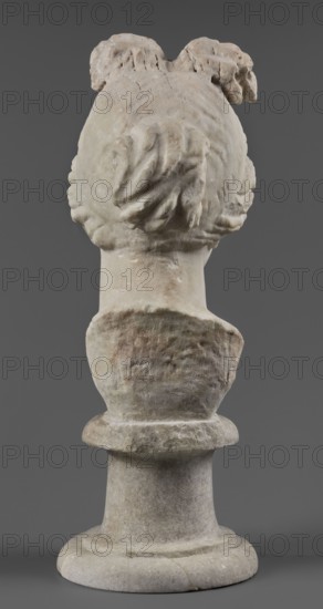 Head of the goddess Aphrodite, Roman workshop, Roman sculpture portrait, early 1st century AD, sculpture; restitution Przeworsk; Aphrodite mythology., The image shows the rear view of a weathered stone bust on a round pedestal against a plain dark gray background. The bust has textured, carved hair gathered at the back with visible waves and folds, and the stone surface displays uneven wear and pitting. The pedestal and lower portion of the bust share the same mottled stone appearance with areas of lighter and darker beige, off-white, pale gray, and subtle brownish tones. The overall color palette includes shades of gray, beige, off-white, and light brown.