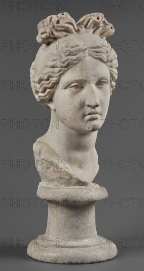 Head of the goddess Aphrodite, Roman workshop, Roman sculpture, portraits, early 1st century 1st century AD, sculpture, Aphrodite mythology, A marble bust of a person shown from the shoulders up on a round pedestal, carved with wavy hair pulled back into a gathered topknot, a smooth forehead and calm facial features including closed eyes and a neutral mouth, with visible surface wear and small chips on the nose, chin, and hair; the sculpture displays tones of off-white, beige, and light gray against a solid gray background.
