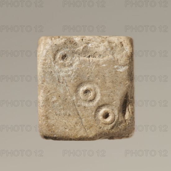 Six-sided dice with pips 1–6 shown as hollow circles and dots; material unspecified; maker and place of origin, height 0.9 cm, width 1.1 cm, depth 0.9 cm., A roughly cubic object with a worn, textured surface in shades of beige, tan, and light brown, showing three recessed circular depressions on one face and slight chips and abrasions along the edges, set against a neutral light gray-beige background.