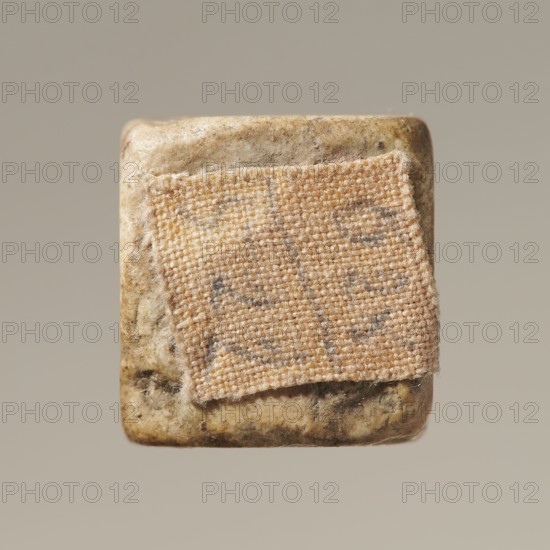 Dice, six-sided dice for the game of dice, points 1–6 shown as hollow circles and dots, material unspecified, place of origin unknown, varia, height 0.9 cm, width 1.1 cm, depth 0.9 cm, A close-up of a square object with rounded corners showing a textured fabric patch adhered to its surface; the object surface is mottled with beige, tan, brown, and gray tones, and the fabric patch is woven with visible fibers in shades of light tan and beige with subtle gray flecks, frayed edges, and a slightly raised, coarse texture against the smoother, speckled background.