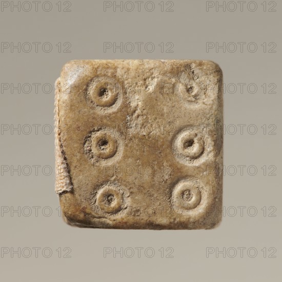 Dice, six-sided dice with pips 1–6 represented as hollow circles and dots, unspecified origin, varia; height 0.9 cm, width 1.1 cm, depth 0.9 cm., A square, worn object with rounded corners and a rough, textured surface in shades of beige, tan, and light brown, showing a pattern of a few circular depressions with raised rims on one face; a small strip of frayed fabric or cord in a pale beige color is attached along one edge; the background is a smooth, muted grayish-beige.