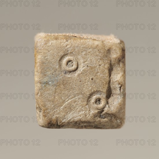 Dice, dice game. Six-sided dice with pips 1–6 shown as hollow circles and dots. Material unspecified., Varia. Height 0.9 cm; width 1.1 cm; depth 0.9 cm., A close-up of a small, roughly cube-shaped object with a weathered, pitted surface in beige and tan tones with hints of light brown and faint bluish-gray smudges; the visible face shows a few shallow circular impressions and irregular indentations, with worn rounded edges and subtle surface scratches, set against a plain neutral beige background.
