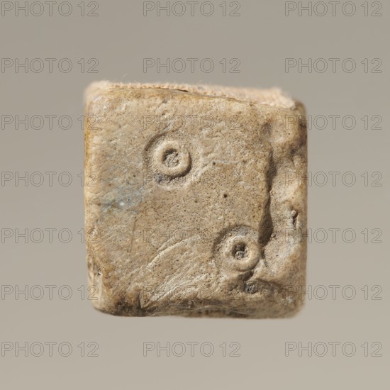 Six-sided dice with pips 1–6 shown as hollow circles and dots; material unspecified; maker and place of origin, height 0.9 cm, width 1.1 cm, depth 0.9 cm., A small, worn cube-like object with a rough, pitted surface in shades of beige, tan, and light brown, showing a few circular depressions on its visible face and irregular edges with slight darker brown discoloration and faint traces of gray-blue; the background is a uniform muted gray-beige.
