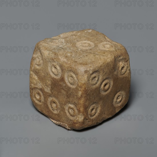 Dice, six-sided dice with pips 1–6 shown as hollow circles and dots, unspecified origin, varia, height 0.9 cm, width 1.1 cm, depth 0.9 cm, A roughly cube-shaped stone with rounded edges and an uneven surface sits on a flat background; the stone is predominantly tan and brown with lighter beige patches and subtle darker brown areas, and each visible face is marked by a pattern of shallow, circular depressions arranged in groupings, giving a textured, pitted appearance.