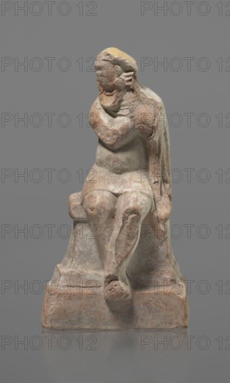 Terracotta figurine of an actor, Terracotta; Hellenistic period, Greece, 332–30 BC. Figurine of an actor sitting on a pedestal. Head turned to the right; legs crossed at the ankles. Wearing a loincloth and a cloak covering the left side of the body. Left arm bent at the elbow, hand rests on the right cheek. Right hand holds the cloak on the left shoulder. Face covered by a caricatured mask with a beard. Sculpture; height 18.2 cm. Preservation: fragmentary—cheek, left hand, and left calf are imprinted., A worn seated statue on a rectangular base, showing a figure with a rounded head and softened facial features, one arm raised toward the face and the other resting at the side, draped cloth falling over the back and beside the body, visible surface erosion and flaking, textured rough patches and smoother areas, and a color palette of earthy beige, pale brown, muted gray, and touches of yellow.