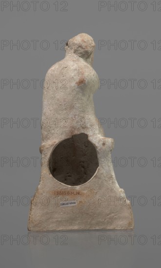 Terracotta figurine of an actor, terracotta, actors, ancient theater, male figurines. Figurine of an actor sitting on a pedestal. The head is turned to the right. The legs are crossed at the ankles. The man is wearing a loincloth and a cloak covering the left side of the body. The left arm is bent at the elbow, and the hand rests on the right cheek. The right hand holds the cloak on the left shoulder. The face is covered by a caricatured mask with a beard. Preservation condition: fragment of the cheek, left hand, and left calf is imprinted. 332–30 BC; Hellenistic period, Greece, sculpture, height 18.2 cm, A weathered sculptural object viewed from the back, showing a rounded upper form resembling a head and shoulders that transitions into a broader base with a large circular hollow in its center; the surface is rough and pitted with an aged, eroded texture. The visible colors include beige, light tan, pale pinkish tones, and patches of brown and gray, with subtle darker shading inside t...