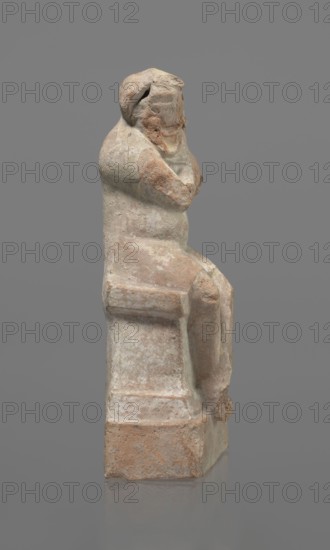 Terracotta figurine of an actor, Terracotta; Hellenistic period, Greece, 332–30 BC. Figurine of an actor sitting on a pedestal. The head is turned to the right. The legs are crossed at the ankles. The man wears a loincloth and a cloak covering the left side of the body. The left arm is bent at the elbow, and the hand rests on the right cheek. The right hand holds the cloak on the left shoulder. The face is a caricatured mask with a beard. Preservation condition: fragmentary—cheek, left hand, and left calf are imprinted. Height 18.2 cm., A seated statue rendered in weathered stone with a muted palette of beige, pale gray, and warm terracotta tones, showing a figure with crossed arms and a rounded head that has eroded facial features; the figure sits on a block-like seat with softened edges and visible surface wear, including patches of lighter and darker discoloration and rough texture; the background is a uniform medium gray.