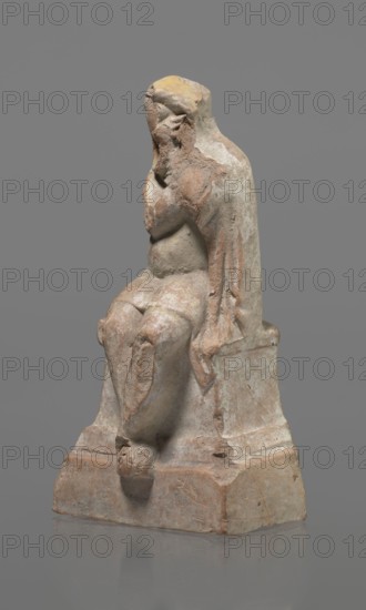 Terracotta figurine of an actor, Terracotta; Hellenistic period, 332–30 BC, Greece. Sculpture, height 18.2 cm. Figurine of an actor sitting on a pedestal. The head is turned to the right. The legs are crossed at the ankles. The man wears a loincloth and a cloak covering the left side of the body. The left arm is bent at the elbow, the hand rests on the right cheek. The right hand holds the cloak on the left shoulder. The face is covered by a caricatured mask with a beard. Preservation: fragmentary — part of the cheek, left hand, and left calf is imprinted., A weathered statue of a seated figure on a block-like base, with a draped garment over the shoulders and back, showing worn facial features and a hand raised toward the face; the surface shows rough, chipped textures and areas of erosion. The colors visible are gray, beige, tan, brown, and patches of yellow.
