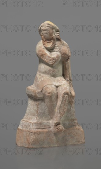 Terracotta figurine of an actor, terracotta, actors, ancient theater, male figurines. Figurine of an actor sitting on a pedestal. The head is turned to the right. The legs are crossed at the ankles. The man is wearing a loincloth and a cloak covering the left side of the body. The left arm is bent at the elbow, and the hand rests on the right cheek. The right hand holds the cloak on the left shoulder. The face is covered by a caricatured mask with a beard. Preservation condition: fragment of the cheek, left hand, and left calf is imprinted. 332–30 BC; Hellenistic period, Greece, sculpture, height 18.2 cm., A weathered stone figure seated on a block-like base, showing a human form with a rounded head, indistinct facial features, one arm bent across the chest and the other resting by the side, legs bent at the knees with one foot projecting forward; the surface is rough and eroded with patches of pale beige, light brown, muted reddish-brown, and traces of greenish-gray, set against a sol...