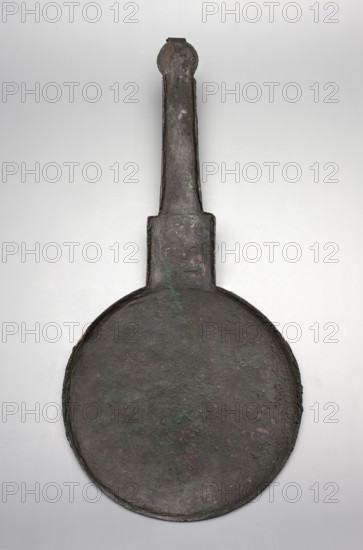 Hand mirror, creator unknown, late 4th century BC, place of origin unknown, mirror, height 36.5 cm, width 17.7 cm, A large round metal object with a long narrow handle attached at the top, both showing a rough, pitted surface and corrosion; the metal appears dark brown to black with areas of green and small patches of pinkish discoloration, set against a pale gray background.