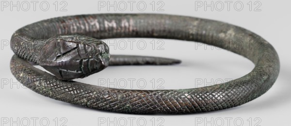 Snake bracelet, snakes, bracelets, 1st century BC, Italy, jewelry; height 2.3 cm, width 9.7 cm, depth 9 cm, weight 174 g, A coiled object shaped like a snake with a sculpted head and textured body rests on a pale surface; the head shows carved features and angular lines while the body displays a crosshatched scale pattern. The object has a metallic appearance with a patina showing dark brown, green, and hints of black and coppery tones. The background is a smooth, light gray, and the lighting highlights the raised texture and subtle color variations on the surface of the coil.