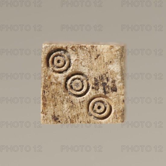 Dice, toys, dice, parlor games, 323 BC–395 AD; Hellenistic period–Roman period, Greece, toy, A small square object with a rough, textured surface in beige and tan tones sits against a plain light gray-beige background; the object shows a pattern of three circular indentations arranged diagonally, each indentation having concentric rings with darker brown residues, and the square surface displays a mottled mix of light beige, tan, brown, and darker specks with visible grain and uneven edges.