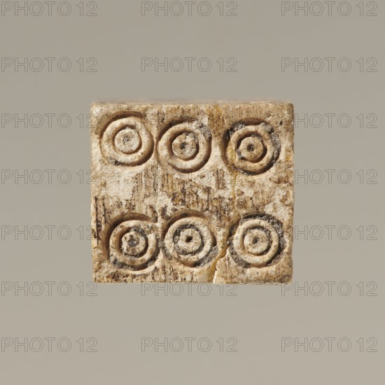 Dice, dice, toys, parlor games, 323 BC–395 Hellenistic period–Roman period, Greece, toy, A square, beige-toned carved object with a rough, weathered surface showing a pattern of concentric circular grooves arranged in a grid; the object displays variations of beige, tan, cream, brown, and hints of darker gray in recessed areas, with visible texture, small pits, and some surface discoloration against a muted light gray-beige background.