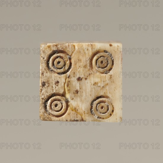 Dice, dice, toys, parlor games; 323 BC–395 Hellenistic period–Roman period; dating previously Hellenistic period, Greece; toy, A small square object with a worn, creamy ivory surface showing darker brown spots and stains, featuring a pattern of concentric circular impressions arranged in a grid; the object has visible cracks and rough edges with areas of yellowish discoloration and darker grime, set against a plain soft gray-beige background.