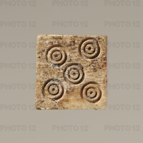 Dice, dice, toys, parlor games, 323 BC–395 Hellenistic period–Roman period, Greece, toy, A square, weathered object with a rough, textured surface in beige and light tan tones, showing circular carved depressions arranged in a scattered pattern; the carved circles have darker brown and grayened interiors and edges, with some areas showing subtle reddish discoloration along one side and small dark specks and stains across the surface; the background is a uniform muted gray-beige.