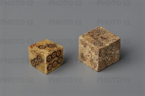 Dice, toys, dice, parlor games, 323 BC–395 AD; Hellenistic period–Roman period, Greece; toy, A pair of worn cube-shaped objects sit on a smooth surface, one smaller and the other larger, both showing a mottled, aged beige and brown coloration with darker circular indentations on their faces; the cubes display textured, scuffed surfaces with areas of darker brown staining and small flecks of reddish-brown, while the background and surface are a consistent mid-gray.