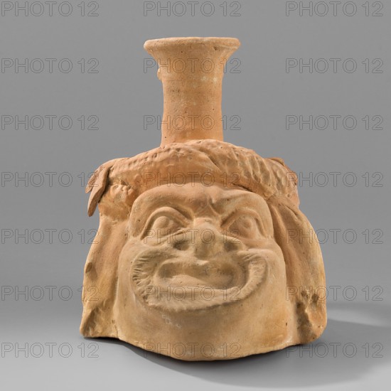 Vessel with a belly in the shape of a comic mask; unknown Greek workshop; plastic vessel; theatrical mask motif; Hellenistic period, 330–30 BC; Greece : vessel; height 10.6 cm, width 9 cm, depth 11.7 cm, weight 0.210 kg., A terracotta sculpted head with a flared neck rising from the top, rendered in a warm orange-brown clay color, sits against a smooth grey background. The face has exaggerated features with deep-set eye shapes, a broad flattened nose, pronounced cheek contours, and a wide open mouth showing sculpted lips and textured inner mouth. A textured band resembling hair or a headdress runs across the forehead with a few raised ridges and a small leaf-like protrusion at one side. The surface shows variations in the orange-brown tone with some lighter and slightly darker patches and a few small surface imperfections. The background is uniformly grey and smooth.