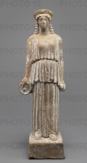 Female terracotta figurine of a standing woman positioned frontally on a quadrangular base. The figure's head, set on a massive neck, is adorned with a polos diadem. The face is oval with a large nose, parted mouth, and clearly defined eyebrow arches. Hair, pinned under the polos, falls over the shoulders and back. The body is almost entirely covered by a peplos with a long apotygm tied at the waist; the garment falls to the ground creating regular, horizontal folds. Arms fall along the torso; in her right hand she holds a patera, and in her left she clutches an object resembling an apple or pomegranate the latter supports an identification as Persephone. Traces of a chalk polychrome base are visible across the surface; remnants of light red paint are visible in the recesses of the robe and remnants of dark red polychrome on the base. Ca. 450 BC, Attica. Figurine; height 34 cm, width 10.5 cm, depth 9 cm, weight 0.75 kg., A carved standing figure of a person on a rectangular base, rende...