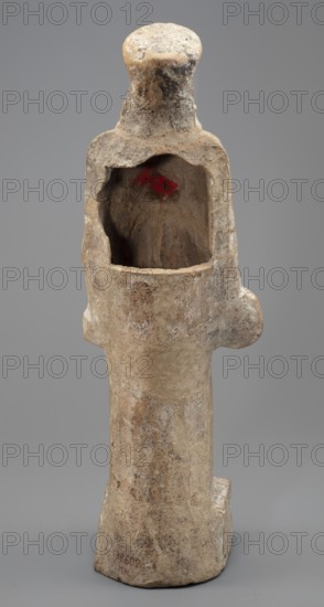 Female terracotta figurine of a standing woman positioned frontally on a quadrangular base. The figure's head, set on a massive neck, is adorned with a polos diadem. The face is oval with a large nose, parted mouth, and clearly defined eyebrow arches. Hair is pinned under the polos and falls over the shoulders and back. The body is almost entirely covered by a peplos with a long apotygm tied at the waist; the garment falls to the ground creating regular horizontal folds. The arms fall along the torso; in the right hand she holds a patera, in the left an object resembling an apple or pomegranate—this attribute allows identification as Persephone. Traces of a chalk polychrome base are visible across the surface; remnants of light red paint remain in the robe’s recesses and dark red polychrome on the top of the base. Ca. 450 BC, Attica. Figurine. Height 34 cm, width 10.5 cm, depth 9 cm, weight 0.75 kg., A weathered vertical stone figure with a rounded top and a hollowed rectangular cavity...