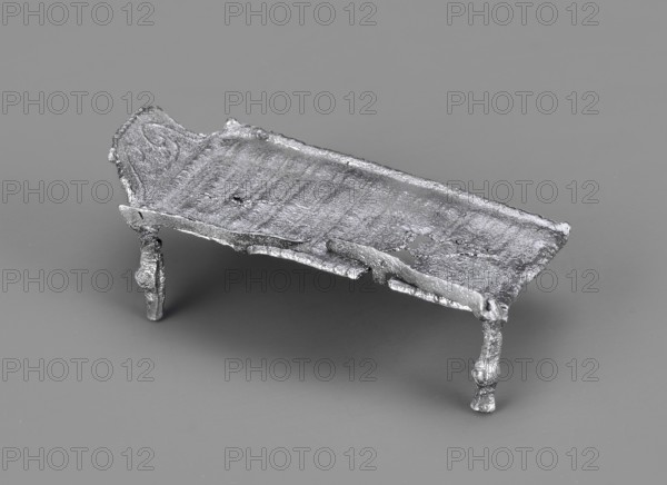 Miniature bed, votive offering or model of furniture; rests on four legs formed from square bars with spherical bulges at mid-height. The bed plate, covered with parallel slightly convex lines arranged crosswise, has long edges terminating in inwardly curled cylinders; shorter edges decorated with a row of small convex lines. A railing is attached to one long edge, mounted on four equidistant posts: two in the corners and two between them. Inside the headboard is a plastic decoration of two diagonally arranged volutes growing from the lower corners, highlighted by an arched line. 1st–2nd century Imperial Period, Roman Empire 27 BC–476, function: toy or amulet. Dimensions: height 2.1 cm, width 5.9 cm, depth 3 cm; weight 0.017 kg., A small metallic object resembling a low rectangular platform with rough, uneven surfaces and irregular edges sits on a smooth background. The platform has a textured top with visible ridges and pitted areas, and a raised section at one short end that appears ...