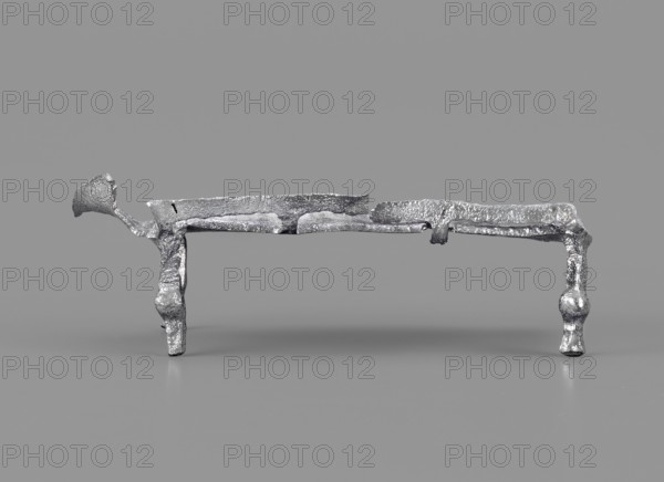 Miniature bed, votive offering or model of furniture; rests on four legs formed from square bars with spherical bulges at mid-height. The bed plate, covered with parallel slightly convex lines arranged crosswise, has long edges terminating in inwardly curled cylinders; shorter edges decorated with a row of small convex lines. A railing is attached to one long edge, mounted on four equidistant posts: two in the corners and two between them. Inside the headboard is a plastic decoration of two diagonally arranged volutes growing from the lower corners, highlighted by an arched line. 1st–2nd century Imperial Period, Roman Empire 27 BC–476, function: toy or amulet. Dimensions: height 2.1 cm, width 5.9 cm, depth 3 cm; weight 0.017 kg., A metallic, silver-colored object with a rough, irregular texture sits on a smooth gray surface against a gray background; the object has an elongated horizontal section supported by a pair of uneven vertical legs, with one end featuring a curved, flared exten...