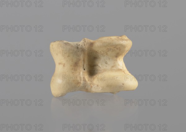 Goat astragal, Myrmekion Crimea, astragal goat condylo-talus bone, Crimea Ukraine, bones, antiquity; 332–30 BC, Hellenistic period, Myrmekion place of finding, product of nature, length 2.1 cm, A single irregularly shaped pale beige object with smooth, rounded surfaces and subtle brownish spots sits against a uniform gray background, with a soft reflection visible on the surface beneath it; the object shows areas of lighter creamy tones, slightly darker tan patches, and tiny darker speckles, and its form includes concave and convex curves with a central indentation.