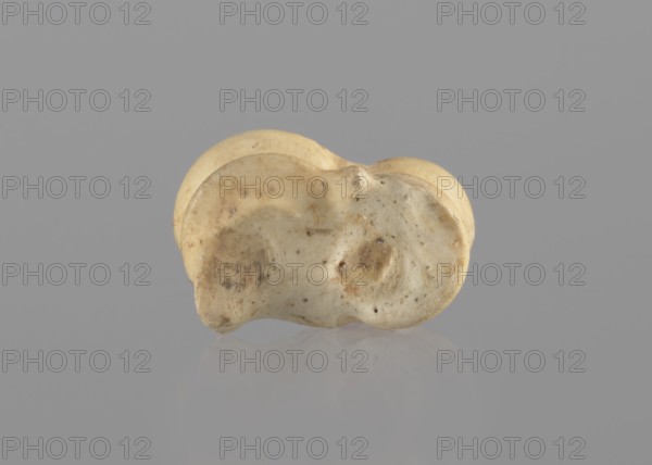 Goat astragal, Myrmekion Crimea, astragal goat condylo-talus bone, Crimea Ukraine, bones, antiquity; 332–30 BC, Hellenistic period, Myrmekion place of finding, product of nature, length 2.1 cm, A small, irregularly shaped object with a smooth, rounded form sits centered on a flat surface against a plain background; the object shows a mix of pale beige, creamy off-white, light tan, and faint brown discoloration with some darker speckles and patches, and it has subtle variations in texture including smoother shiny areas and slightly rougher, pitted spots, with a soft reflection visible on the surface below.