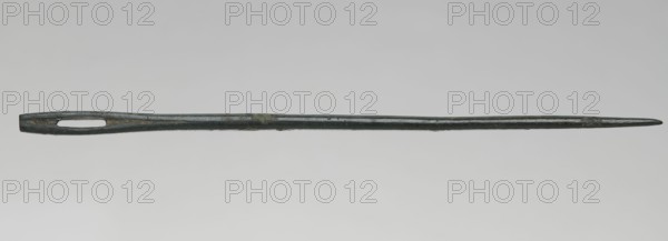 Needle, maker, creator/author entries note Madeyski, Antoni 1862–1939; tool; weight 10 g; height 14.2 cm; diameter 0.4 cm., A long slender object with a tapered pointed end and a wider opposite end featuring an elongated oval opening, shown horizontally against a neutral light gray background; the object appears metallic with a dark greenish-gray patina and small patches of darker and lighter greenish discoloration along its length.
