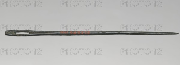 Needle, maker, Madeyski, Antoni 1862–1939 associated; tool; weight 10 g; height 14.2 cm; diameter 0.4 cm., A long, slender metal object with a dark greenish-gray patina stretching horizontally across a pale gray background, featuring a widened end with an elongated oval opening and tapering to a pointed tip at the opposite end; the surface shows areas of darker and lighter green and gray with small patches of brownish discoloration.