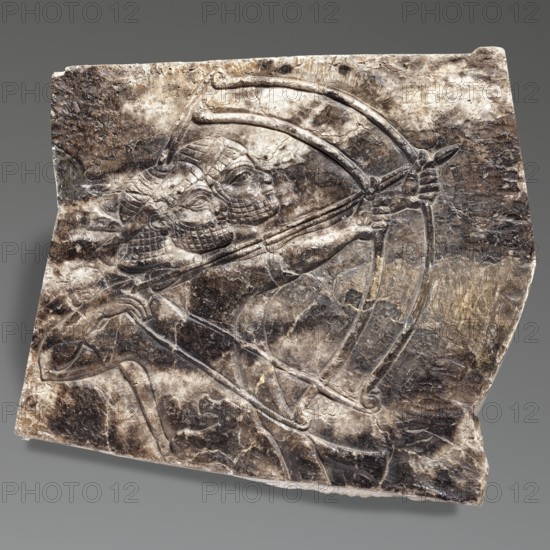 Fragment of an orthostat with relief decoration, unknown Neo-Assyrian workshop. Fragment of a stone wall-facing slab orthostat with a relief representation of two men shooting bows. The man in the foreground holds a bow in his outstretched left hand, drawing it with his bent right hand. The second archer is largely obscured by the first; his head, left side, and left arm holding a drawn bow and arrow are visible. Both archers wear short-sleeved tunics. Heads shown in right profile with identical beard and hairstyle: thick beards depicted by schematic curls arranged in vertical and horizontal rows; headbands with two ends protruding at the back; hair above the headband combed in straight strands over the forehead and falling in thick schematic curls below to the nape. This hairstyle with a headband is characteristic of depictions of Elamite soldiers in Assyrian art. Neo-Assyrian art, 7th century BC, c. 668–c. 631 BC reign of Ashurbanipal. From Kuyunjik Southwestern Palace of Sennacherib...