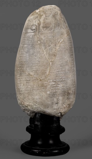 Copy of a Babylonian kudurru from the period of Marduk-naddin-ahhe 11th century BC, Ancient Near East, kudurru copy, Kassite Kingdom 1595–1158 BC, Eastern Gallic scenography, Babylonia Iraq place of origin of the original, 19th century, copy, weight 5.44 kg, A vertically oriented, weathered stone object with an oval top and rounded base sits on a dark, glossy stand; the stone is primarily pale beige with areas of off-white and light brown discoloration and a few darker brown stains, and its surface shows carved, shallow incisions arranged in rows across much of the front; the upper portion of the stone features low-relief carved figures and shapes with similar pale beige and off-white tones; the base of the stone is rougher in texture with more pronounced wear and small chips, and the stand beneath is black and slightly reflective.