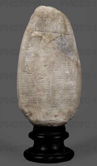 Copy of a Babylonian kudurru from the period of Marduk-naddin-ahhe 11th century BC, Ancient East, kudurru copy, Kassite Kingdom 1595–1158 BC, Eastern Gallic scenography, Babylonia Iraq applies to the original, 19th century, copy, weight 5.44 kg, A tall oval stone object with a rough, light beige surface mounted on a dark black rounded stand; the stone shows incised linear patterns and shallow relief carvings concentrated toward the upper portion while the lower portion displays dense, fine incisions covering much of its face; the top area includes carved architectural and figurative shapes in low relief with worn edges, and the stone has areas of discoloration in slightly darker beige and pale gray; the stand is glossy black with rounded tiers and visible wear marks, and the background is a uniform medium gray.