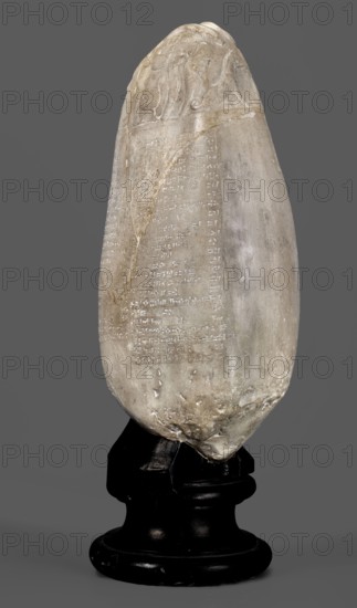Copy of a Babylonian kudurru from the period of Marduk-naddin-ahhe 11th century BC, Ancient Near East, kudurru copy, Kassite Kingdom 1595–1158 BC, Eastern Gallic scenography, Babylonia Iraq applies to the original, 19th century, copy, weight 5.44 kg, A tall, elongated stone object with a rounded top and tapered bottom rests on a black pedestal; the stone is predominantly pale gray with areas of off-white and light beige, showing a weathered, slightly rough surface with smoother patches and faint darker streaks. The top portion has carved relief shapes, and the main body displays a rectangular panel of small incised marks. The lower portion of the stone has irregular chips and a slightly darker, rougher texture where it meets the pedestal, which is glossy black with a turned, rounded base and a few lighter scuff marks. The background is a uniform medium gray.