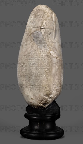 Copy of a Babylonian kudurru from the period of Marduk-naddin-ahhe 11th century BC, Ancient Near East, kudurru copy, Kassite Kingdom 1595–1158 BC, Eastern Gallic scenography, Babylonia Iraq applies to the original, 19th century, copy, weight 5.44 kg, A tall, elongated stone object with a rounded top and an irregular, weathered surface sits on a dark, rounded pedestal; the stone is mostly pale beige and off-white with areas of darker gray and brown discoloration and visible scratches and chips, and there are a few shallow carved panels and low-relief motifs near the upper portion while the lower portion shows a textured, pitted area; the pedestal is black with a slightly glossy finish and shows some worn patches and small scuffs; the background is a flat medium gray.