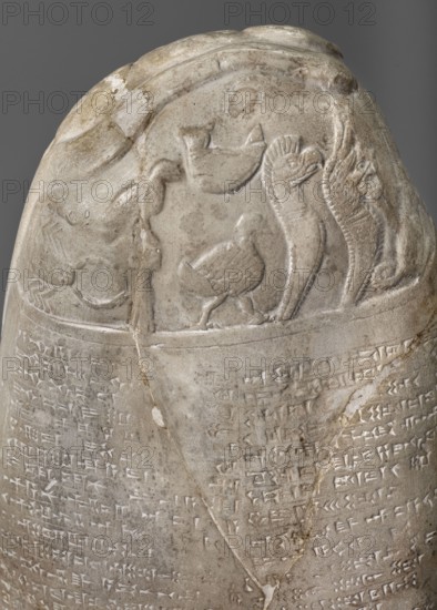 Copy of a Babylonian kudurru from the period of Marduk-naddin-ahhe 11th century BC, Ancient Near East, kudurru copy, Kassite Kingdom 1595–1158 BC, Eastern Gallic scenography, Babylonia Iraq place of origin of the original, 19th century, copy, weight 5.44 kg, A stone object with a rounded top carved in light gray and beige tones, showing a low-relief scene near the top that includes a group of bird-like figures and elongated serpent-like forms, with carved decorative ridges and textured feather details; below the scene the stone surface continues in similar gray and beige hues with a band of closely carved markings forming several horizontal rows, and a visible diagonal crack running through the middle of the object.