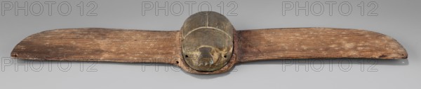 Winged scarab; protective amulet used with mummies and as grave goods; sacred animal motif; scarab amulet, Late Period, Egypt, 664–332 BC; materials not specified; height 5 cm, width 23.8 cm, depth 1.8 cm., A long horizontal wooden object with a central rounded metallic element that resembles a carved head sits on a neutral gray background; the wooden parts show a weathered grain with a medium to dark brown tone and areas of lighter brown wear, while the central rounded piece is a dull greenish-bronze with darker brown and patinated spots; the wood has linear grooves and worn edges, and the metallic section has contoured lines and small holes on either side, with subtle shadows beneath the object.
