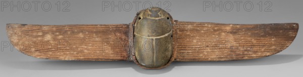 Winged scarab; protective amulet for mummies, wings, grave goods; sacred animal motif; scarab amulet made in Egypt, Late Period, 664–332 BC; materials/technique not specified; dimensions: height 5 cm, width 23.8 cm, depth 1.8 cm., A horizontal wooden object with a central carved beetle-like form attached to a pair of elongated wing-like wooden panels; the central carved form is smooth with rounded contours and shows shades of green and brown with areas of darker patina, while the wing-like panels are textured with visible grain lines and show various tones of brown and tan with lighter dusted spots; the background is a neutral gray and there is a soft shadow beneath the object.
