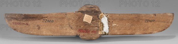 Winged scarab, protective amulet for mummies and grave goods, sacred animal; scarab amulet with wings; Late Period, Egypt, 664–332 BC; height 5 cm, width 23.8 cm, depth 1.8 cm., A worn wooden object with a long horizontal shape and rounded ends rests against a neutral gray background; the wood shows visible grain lines and variations in brown tones, from light tan to medium and darker brown, with some areas appearing slightly reddish-brown and weathered; a central raised section protrudes on the top and bottom, and next to it there is a patch of rough, fibrous material with a whitish residue and a small square of lighter beige material adhered nearby; the surface has scratches, small dark scuffs, and spots of discoloration, and the object casts a soft shadow beneath it.